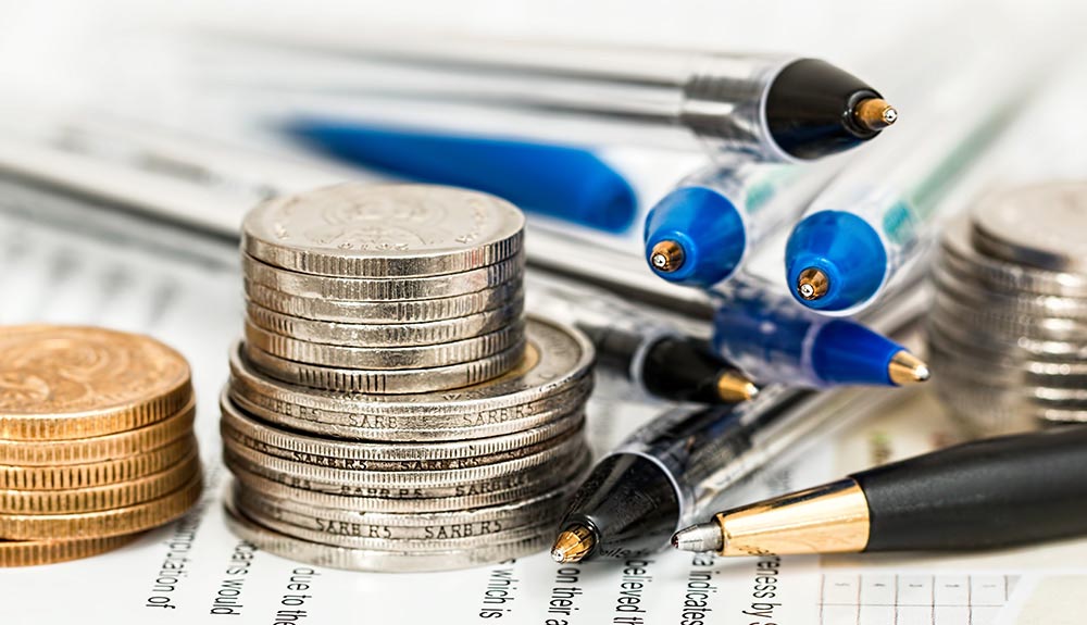 A stack of Canadian loonies, toonies and quarters and a stack of pens sit on a sheet of paper