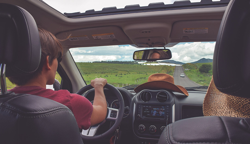 Two people sit in the front of a car, a man in a red shirt driving, his brown cowboy hat sitting on the dash in front of him as he looks out at the rural highway 