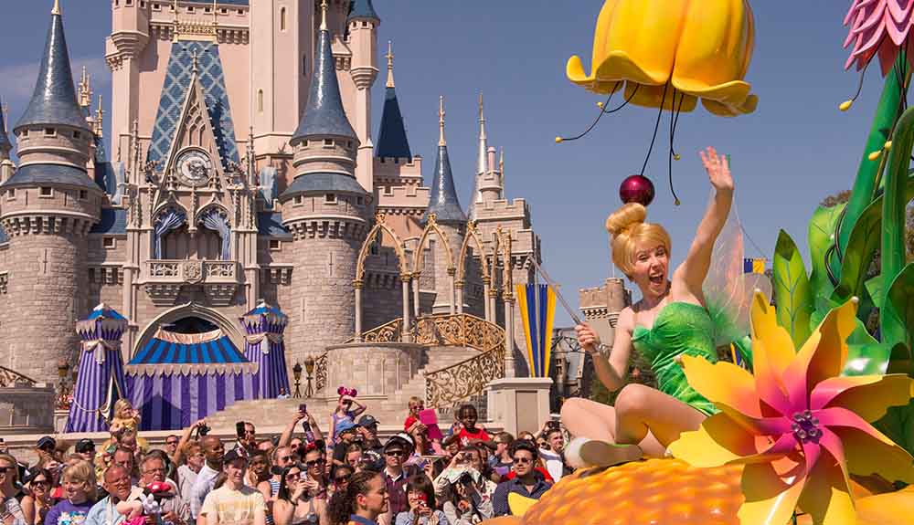 Woman dressed as Tinkerbell waving from a float in a parade as it passes by the entrance to the Magical Kingdom castle