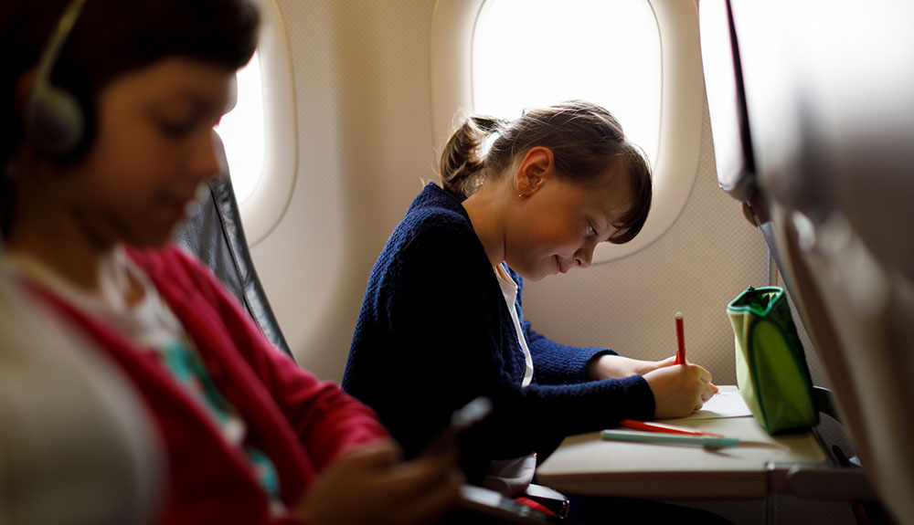 Little girl in her plane seat uses a red writing tool on her tray also tabling her pencil case by a window