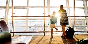 The back of a woman with backpack on the floor and child with backpack from inside the airport looking out as the airplanes