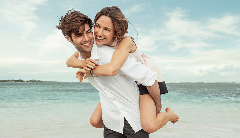 A man gives a piggy-back ride to a brunette woman on the beach