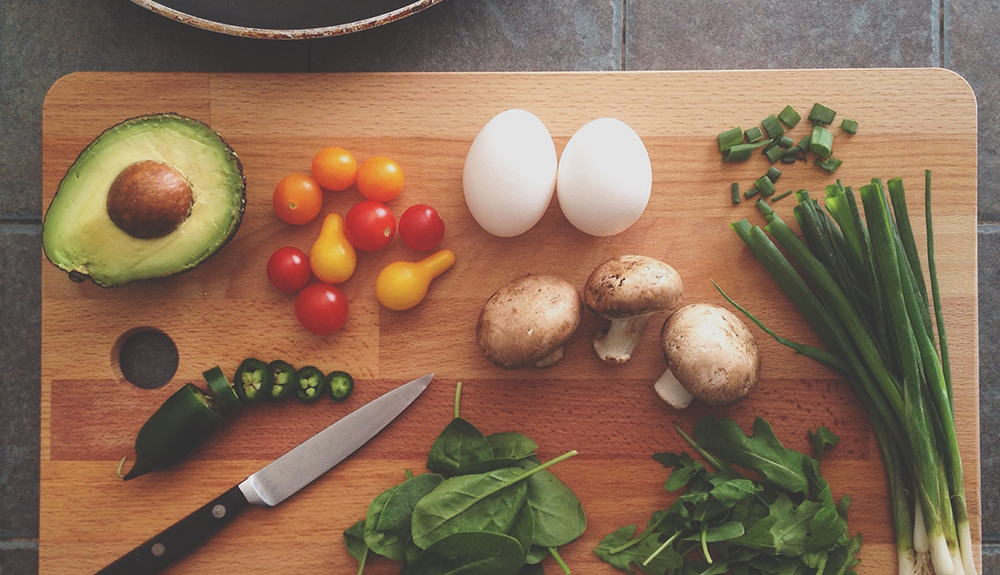 wooden cutting board with knife, half an avocado, jalapeno, tomatoes, eggs, mushrooms, spinach, green onions and arugala