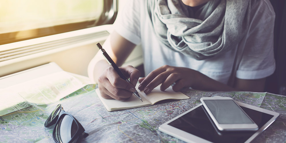 Woman sitting at a table on a train writing into a small notebook on top of a map by her tablet, phone and sunglasses by the window in the daytime 