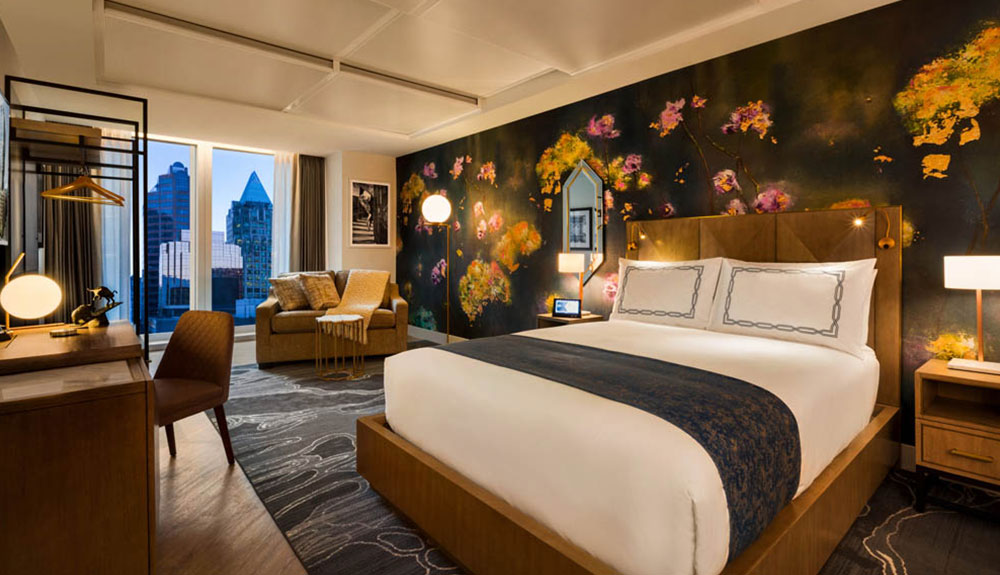 A well-made bed against a floral painted wall, a sofa in the corner by the window and a desk at the foot of the bed in a hotel room