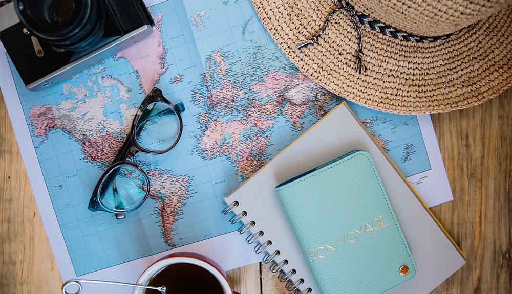 World map on table covered by camera, glasses, sun hat and notebooks