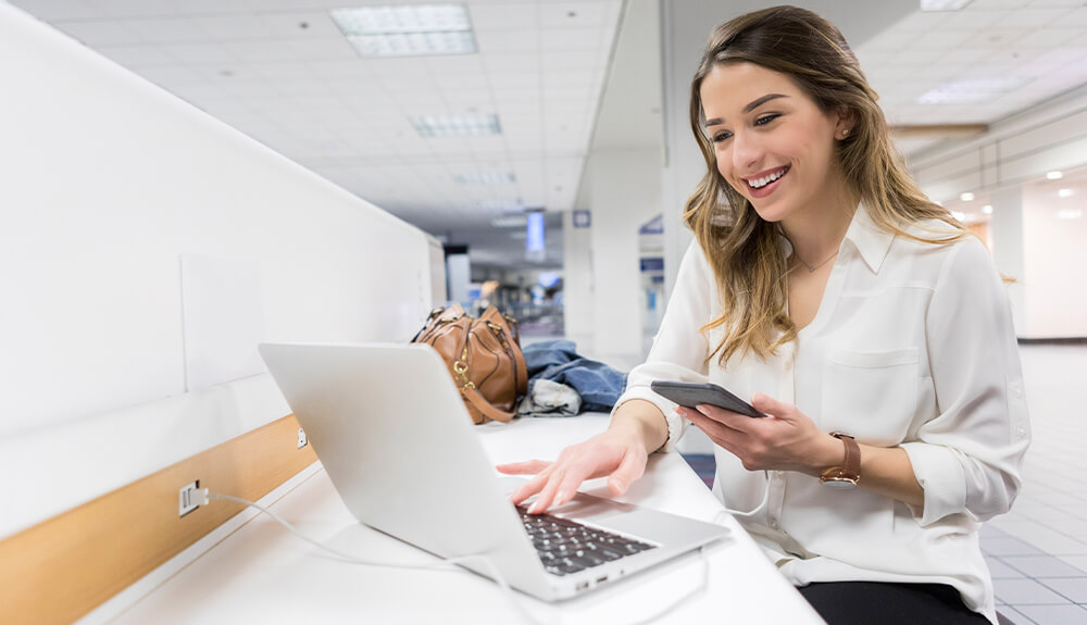 Woman sitting at counter in airport lounge looking at phone and laptop