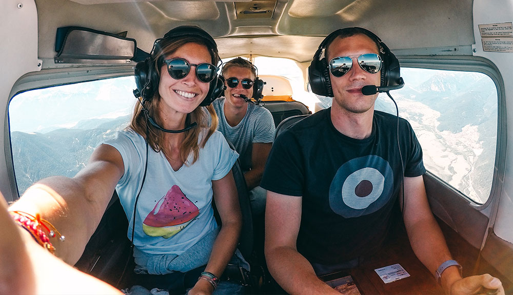 Woman taking the selfie from the front of the plane next to a pilot and another passenger in the back while up in the sky
