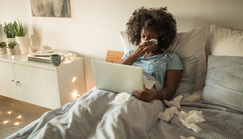 Woman sitting in bed blowing her nose with laptop and pile of tissues next to her