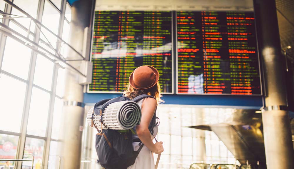 Woman with backpack and rolling suitcase looking at airport departures board
