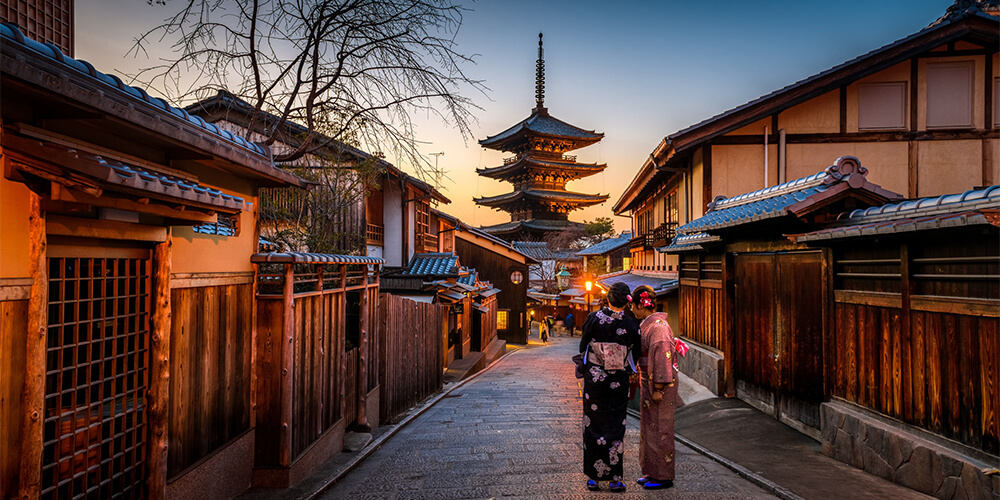 Two women dressed in kimonos walking on street in Kyoto, Japan, lined with traditional buildings at dusk