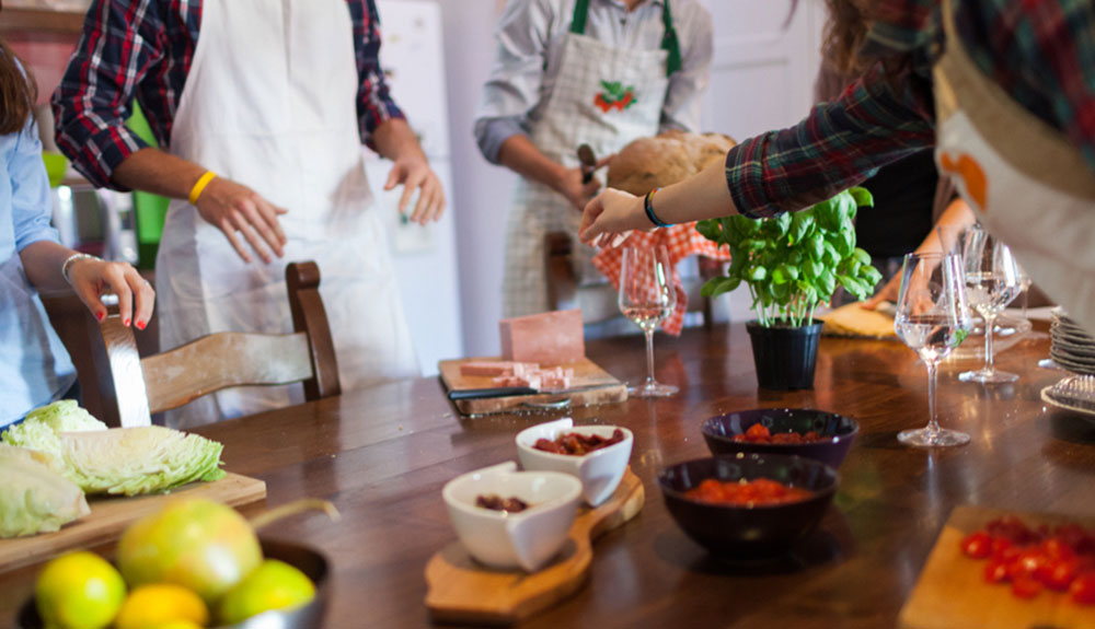 Group of people wearing aprons and hands reaching for the items on the table including lettuce, a bowl of fruit, bowls of cut up tomatoes, glasses, plates and and a plant