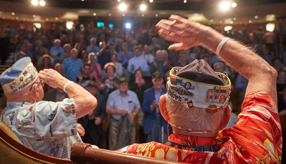 The back of two vets waving to a large audience from a stage