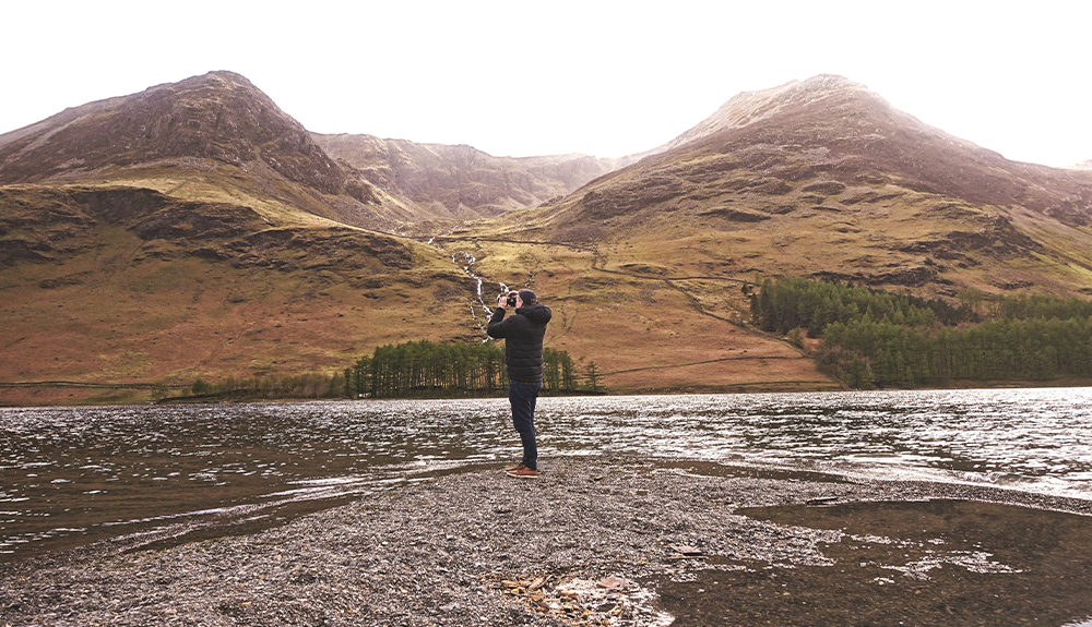 A visitor takes a picture on the shore of a lake beneath a small mountain 