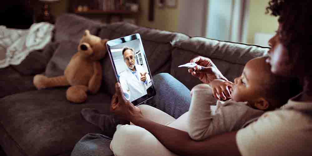 A mother has an onscreen virtual medical consultation with her toddler on her lap