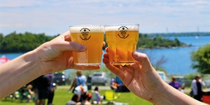 Two people holding glasses of craft beer, clinking the glasses together in a toast, in front of a park on the waterfront.