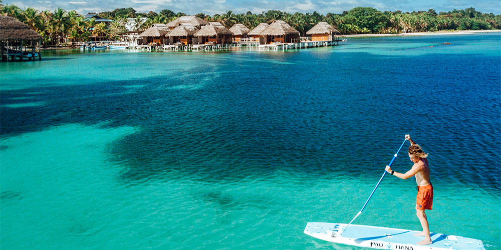 An overhead shot of a person standing on a board out in the ocean, holding a paddle. The ocean is a bright teal colour and in the distance there are bungalows.