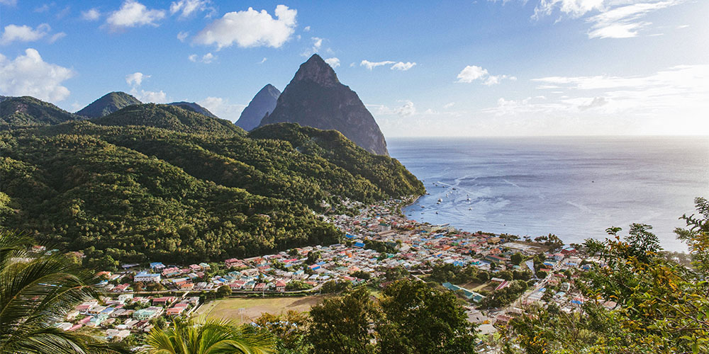 An overhead view of St. Lucia. There are mountains covered in greenery on the left side, and at the bottom centre are differently coloured buildings, and beyond that is the ocean.