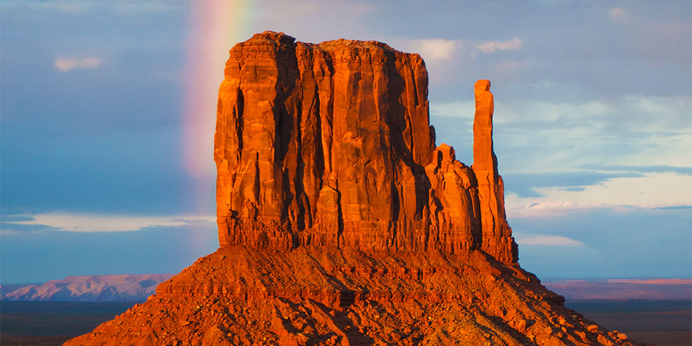A tall, red-orange clay formation at the Grand Canyon, in front of a blue sky and a rainbow.