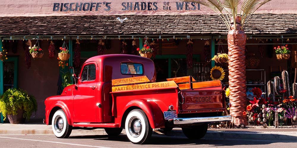 An antique, red pickup truck with tires that have white rims, parked in front of a store.