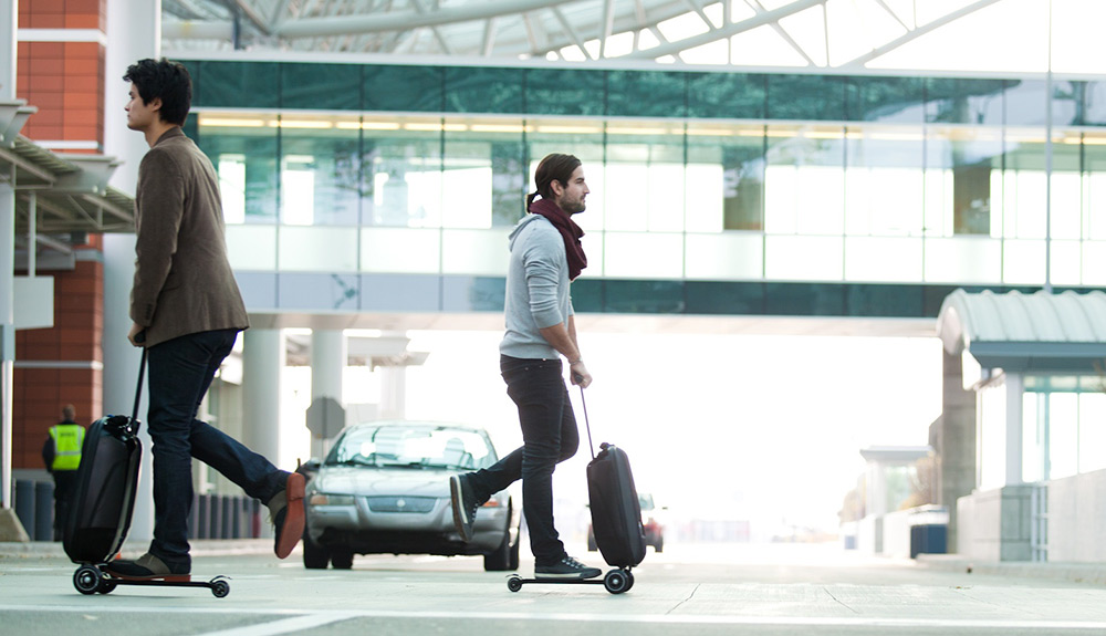Two men are seen riding the Micro Luggage bag that holds out into a miniature scooter
