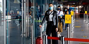 A line of people standing by glass doors. They are each wearing masks with yellow signs next to them indicating that they should keep six feet apart. The woman in front is wearing a cropped black coat  and black pants, holding onto a red rolling suitcase.
