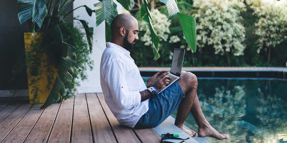 A man wearing a white button up shirt and denim shorts is sitting on the edge of a wooden deck. He is looking at a laptop that’s positioned on his knees, while his bare feet are perched over a turquoise pool. There are lush trees and plants around the pool in the background.