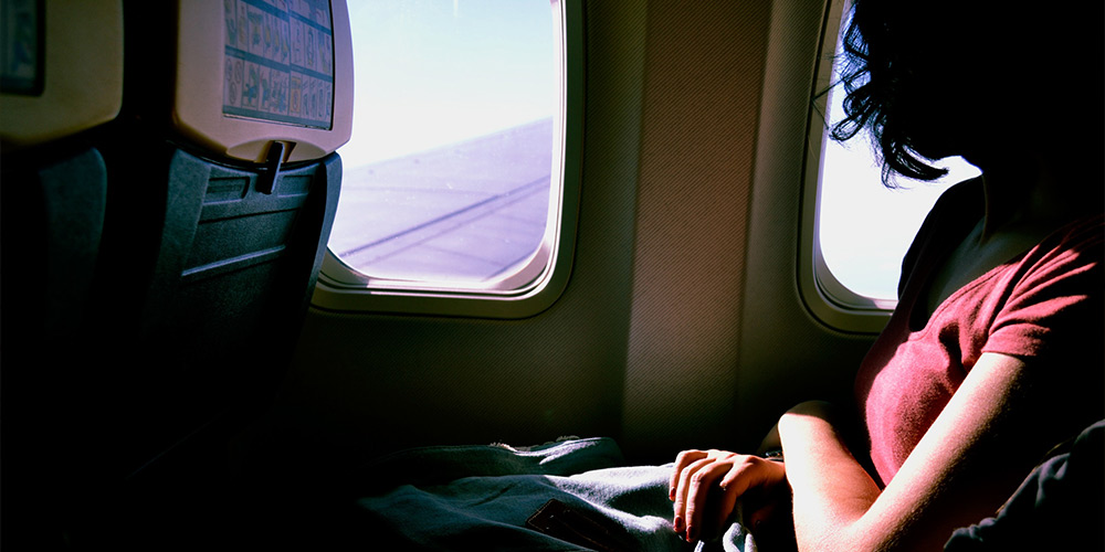 A woman partly obscured by shadows sits with her hands in her lap looking out an airplane window