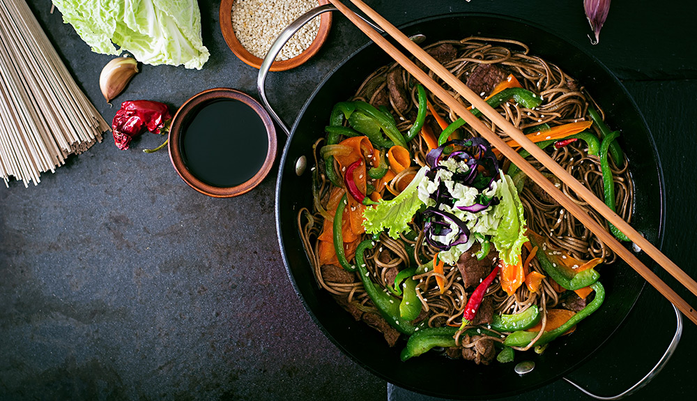 A pot of Soba noodles with peppers, carrots and fresh chillies sits with a pair of wooden chopsticks across it, a small dish with soy sauce and sesame seeds beside it