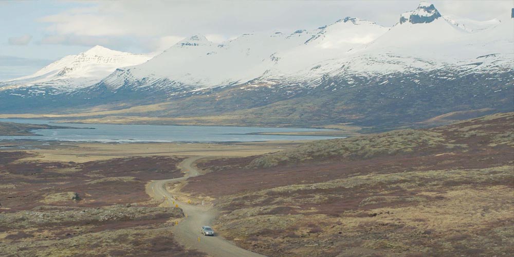 A car drives down a winding road towards snow-covered mountains behind a quiet lake