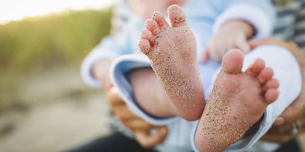 Sandy baby feet seen in the foreground, hands holding baby wearing a light blue playsuit