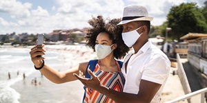 Two people, a man in a white button up shirt and a hat, and a woman in a striped orange, blue and white sleeveless top wearing masks taking a selfie with her cellphone.