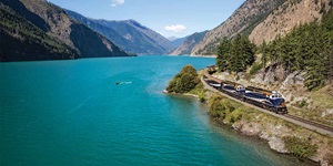 A blue and white train with a wooden roof travels along the tracks on the side of a rocky cliff speckled with green evergreen trees and foliage on the right. There is a large turquoise blue body of water to the left and a mountain range in the distance.  