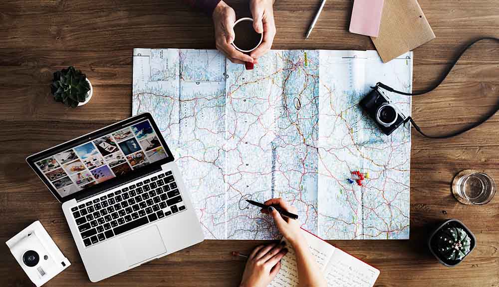 The hands of two people are seen, one cradling a coffee mug, the other with a pen and notebook in hand, pointing to the map that sits between them, a laptop to one side and camera to the other