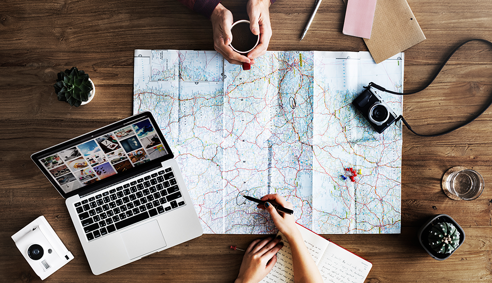 Overhead shot of the hands of two people, one holding a cup of coffee, another pen and notebook in hand pointing to the map that lays between them, a laptop to one side and camera to the other