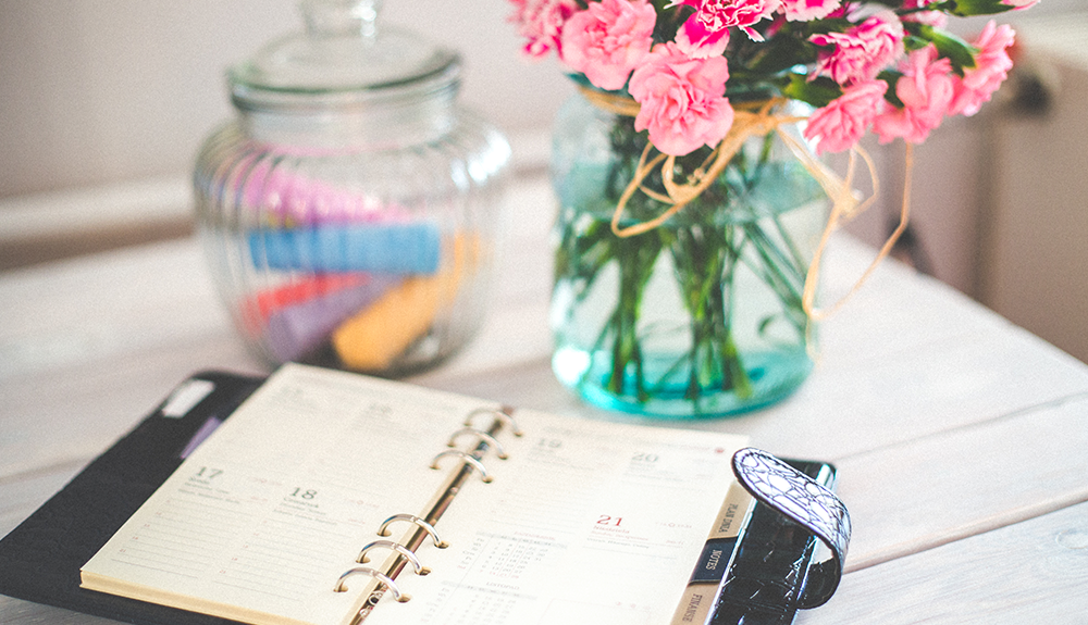 A day planner on a table, a vase of fresh pink flowers behind them
