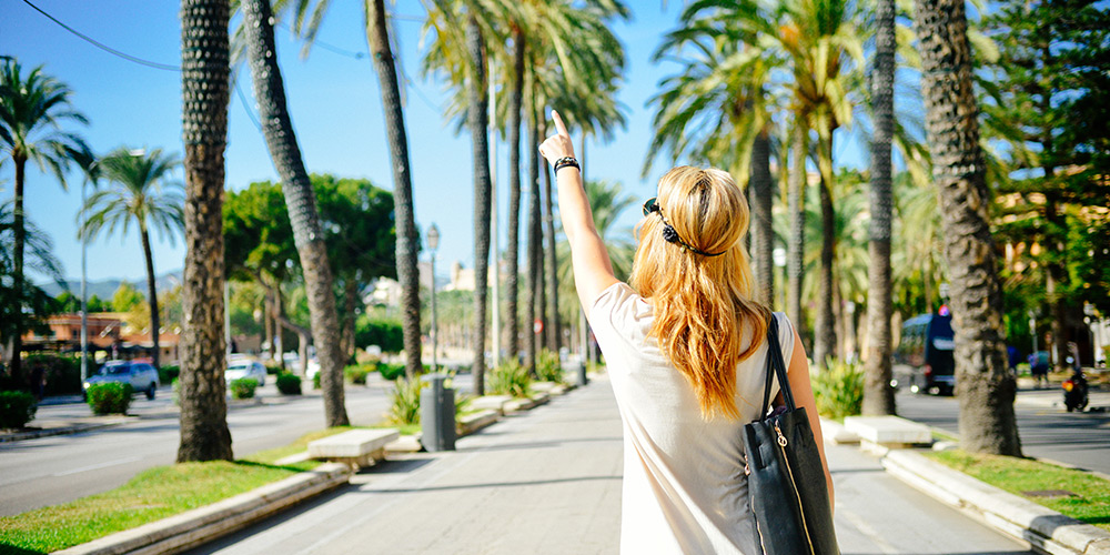 A woman explores a bright street, pointing towards palm trees in the distance