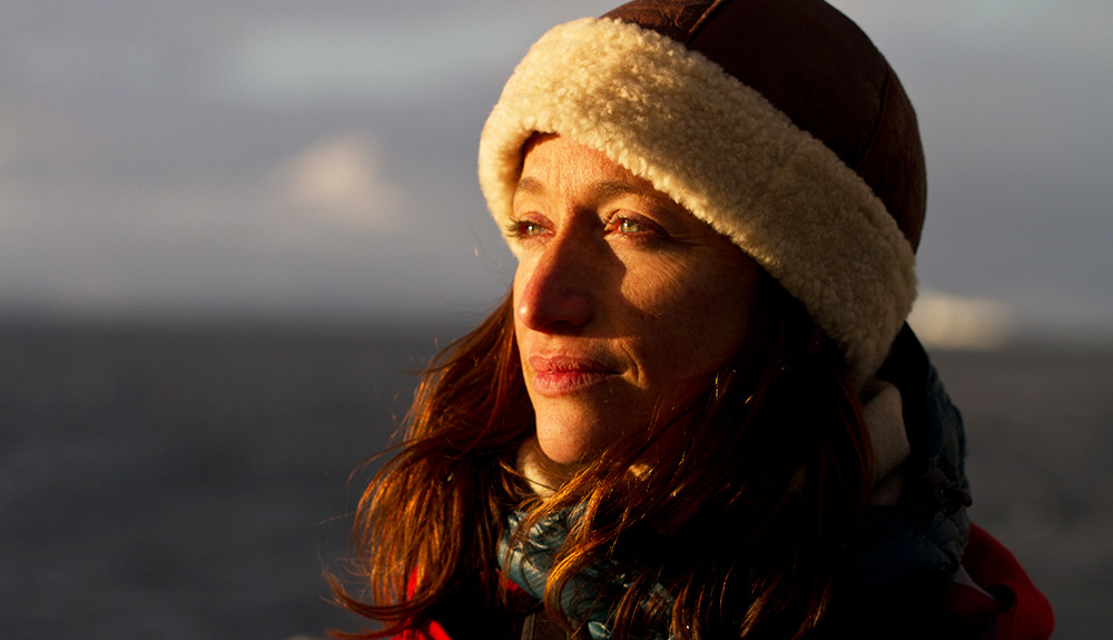 A woman wearing a fuzzy winter headband looks far off in the distance, the setting sun casting a light on her face 