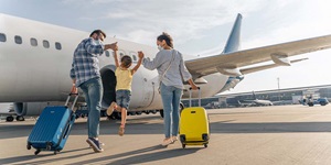 A man wearing a blue plaid shirt and jeans, and a woman wearing a light blue shirt and jeans, each holding onto a rolling suitcase with one hand, are walking on the tarmac towards an airplane. With their other hand, they are each holding onto the hand of a young child who is wearing a yellow shirt and jean shorts. They are swinging her in the air towards the airplane.