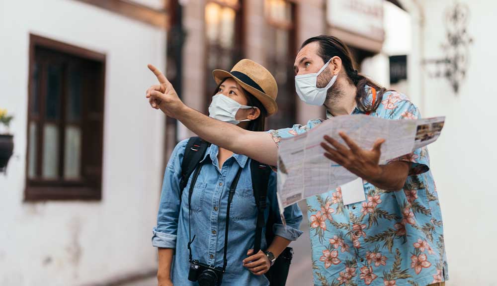 A man wearing a light blue and pink floral shirt and a white face mask is holding a map and pointing at something to the side of him. There is a woman next to him wearing a straw fedora with a black band, a denim shirt and a backpack. She has a camera around her neck and is looking in the direction the man is pointing at.