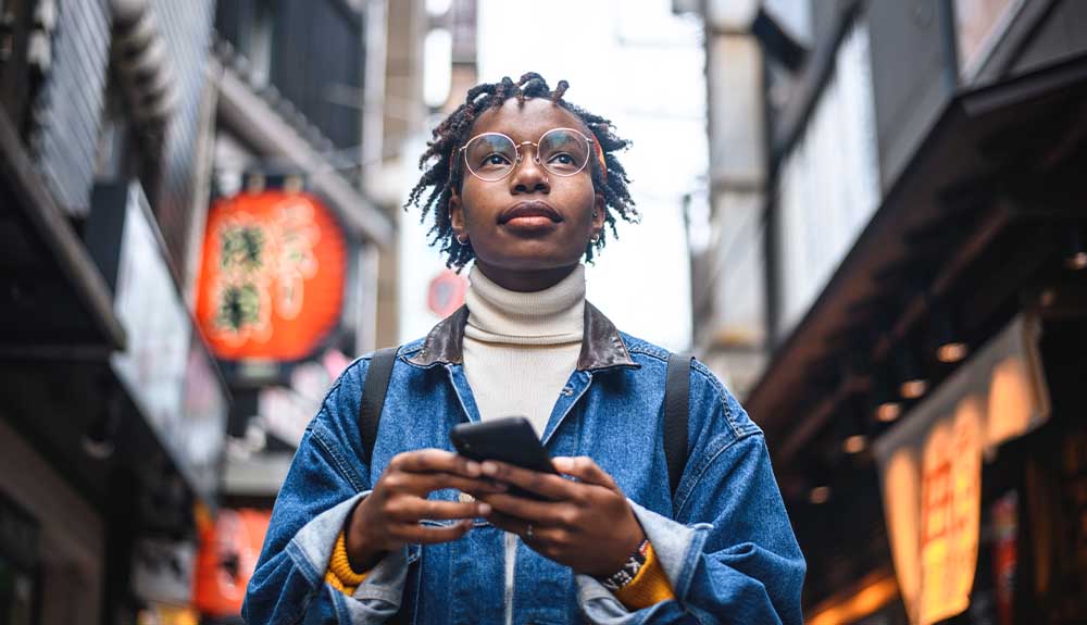 A woman by herself with wire-rimmed glasses wearing a denim coat and white turtleneck holding a cellphone in her hand. Behind her are bright orange lantern-shaped signs in front of storefronts.