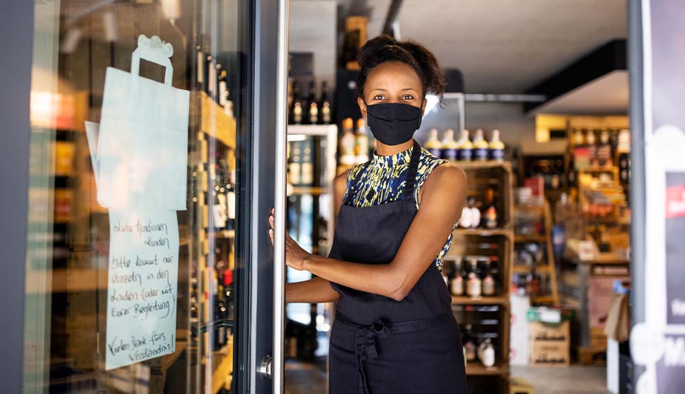 A woman standing at an open store door wearing a black face mask and a black apron. Behind her are wooden shelves stocked with different sized bottles.