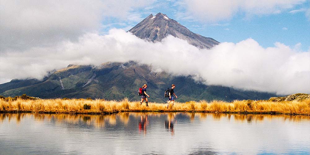 Two people wearing backpacks are walking, one in front of the other. Behind them is a mountain peak and next to them is a body of water.