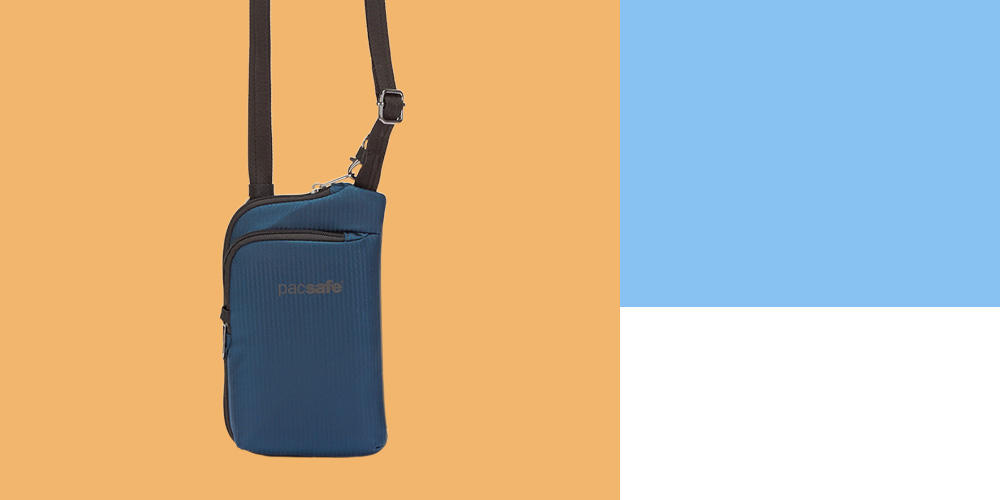 An image of the Pacsafe Daysafe ECONYL® Anti-Theft Tech Crossbody, a blue crossbody bag with a black strap and silver buckle, which says “Pacsafe” on the front, in front of a plain orange background.