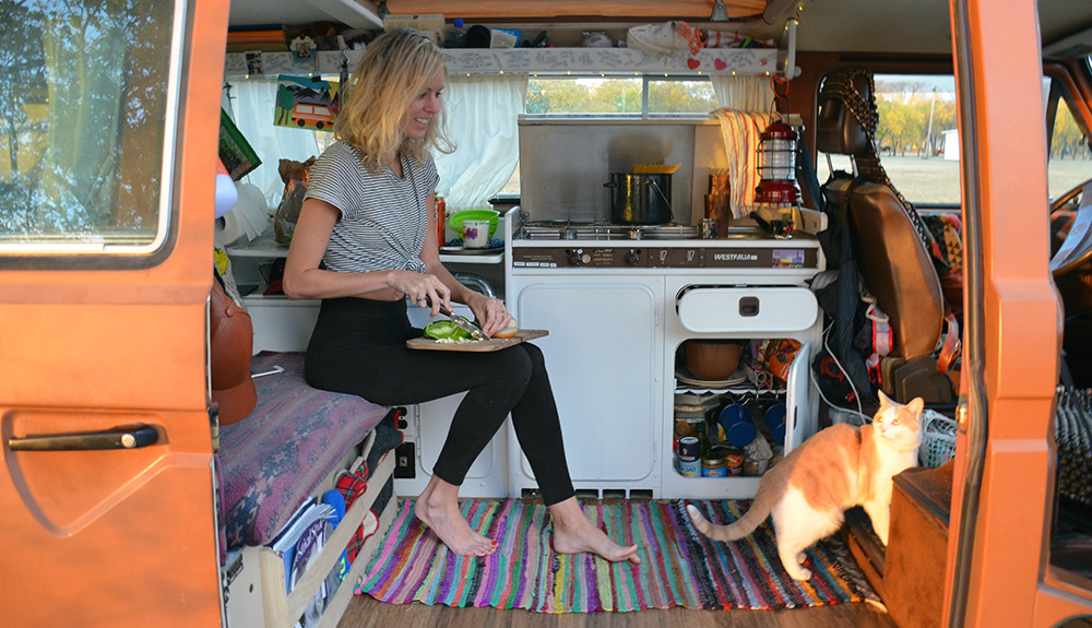 A woman and orange and white cat are seen sitting inside a camper van, the woman preparing a meal and the cat looking back at her