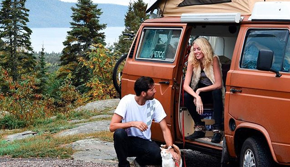A woman sits just inside the door of an orange camper van parked in the wilderness, a man crouches just outside the van petting an orange and white cat on a long red leash