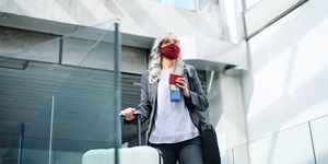 A woman with wavy grey hair wearing a grey suit with a white blouse wearing red framed glasses and a mask. She is holding a blue travel mug in one hand with her tickets. On one shoulder she has a black laptop bag. Her other hand is placed on the handle of a suitcase.