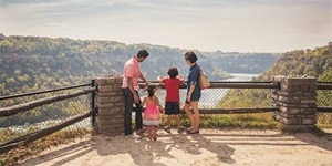 A family are standing at a lookout. There is a brick pillar that is a few feet high with wooden beams forming a fence on either side of it. In front of it is a metal fence. There is a man in a pink button-up short sleeve shirt, jeans and sneakers, a woman in a denim dress with a beige shoulder bag slung over her one shoulder. There are two small kids with them. There is a little girl in a backless pink dress with stripes, and a boy in black shorts, a red T-shirt and sandals. They are all admiring the view in front of them, which is of a river and trees.