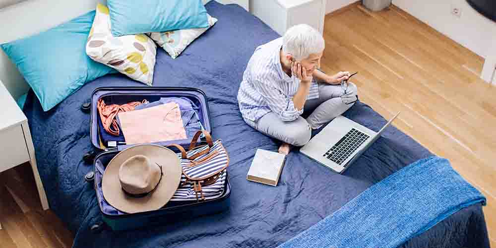 An overhead photo shows a person with grey hair sitting on their bed with a laptop and an open suitcase