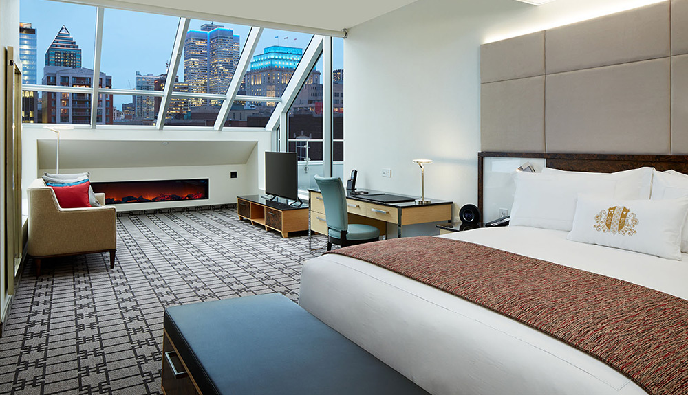 A large hotel room with electric fireplace king sized bed and desk, a skylight looks out at the Montreal skyline